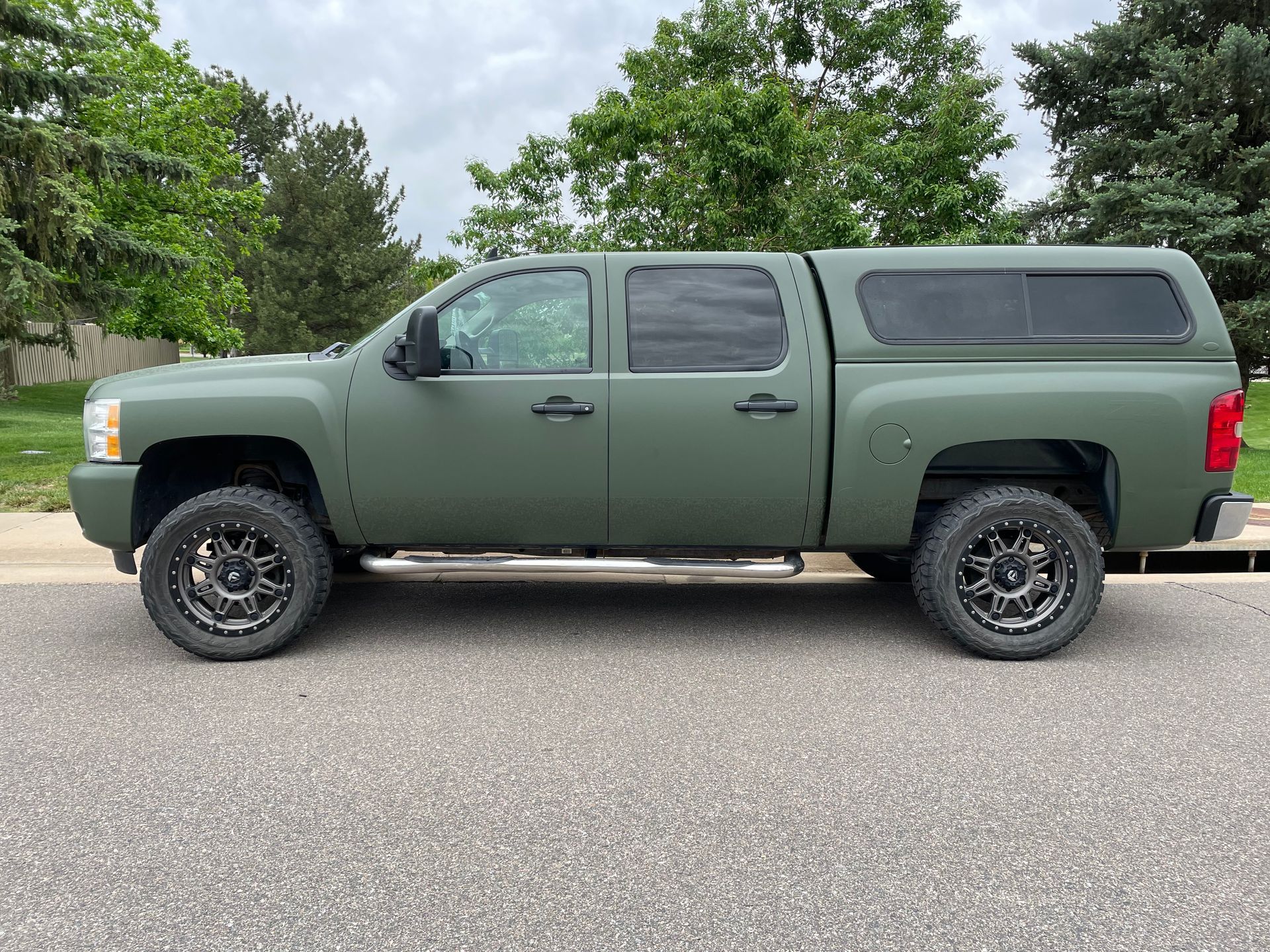 Green pickup truck with camper shell parked on a paved road beside trees