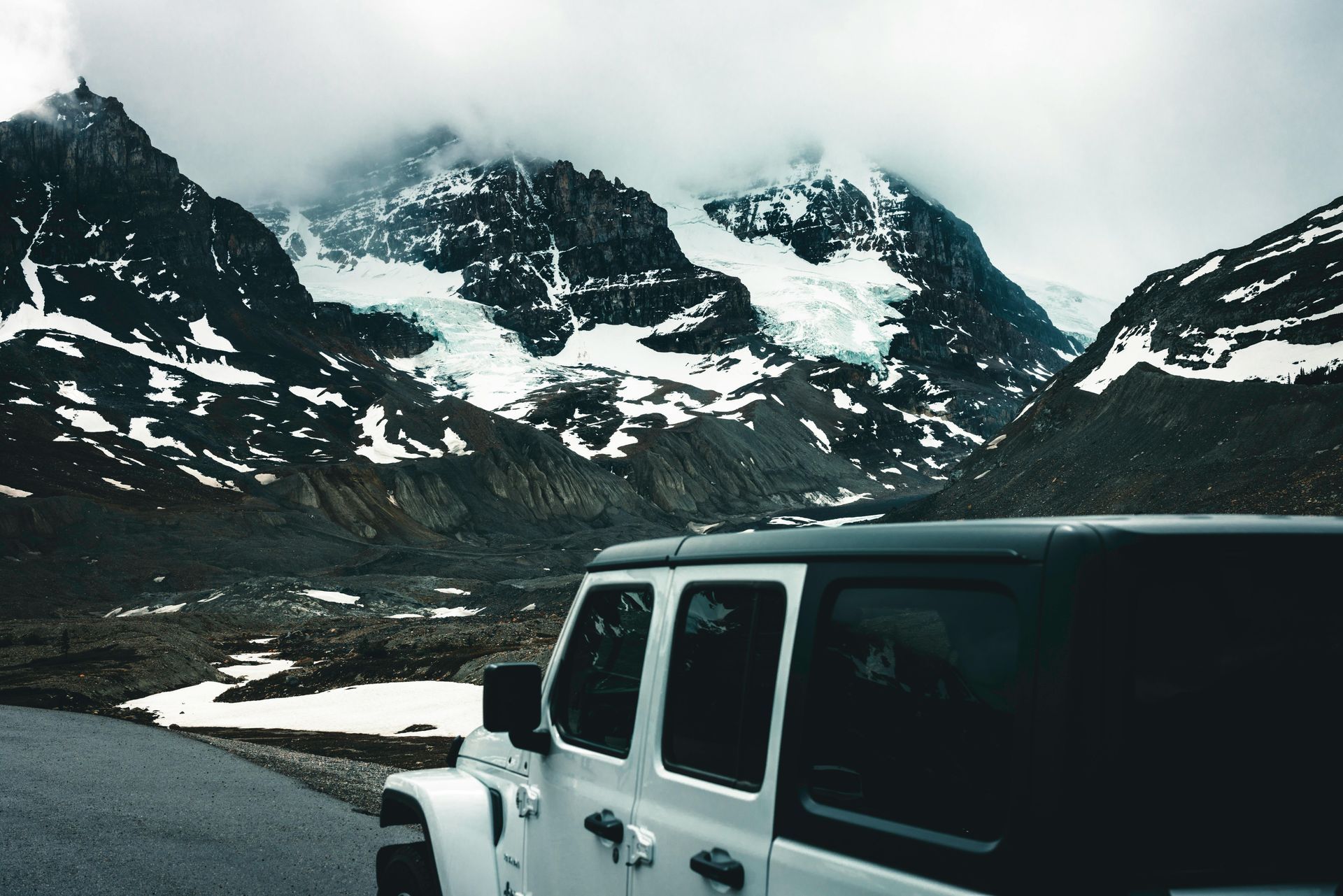 White Jeep parked on a snowy road with rugged mountain peaks under cloudy skies