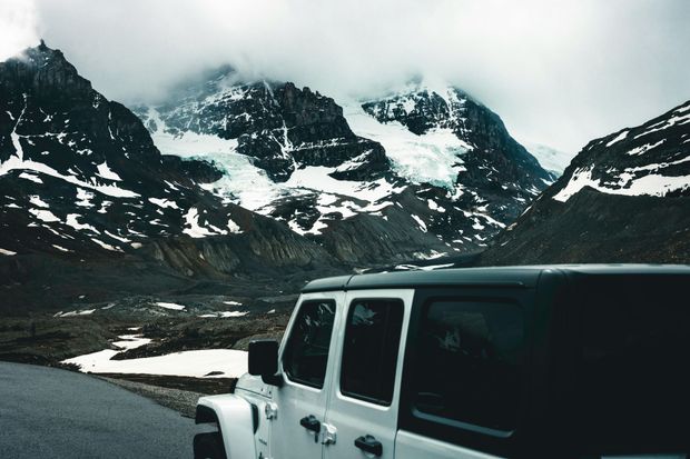 White Jeep on a mountain road with snowy peaks and cloudy sky in the background