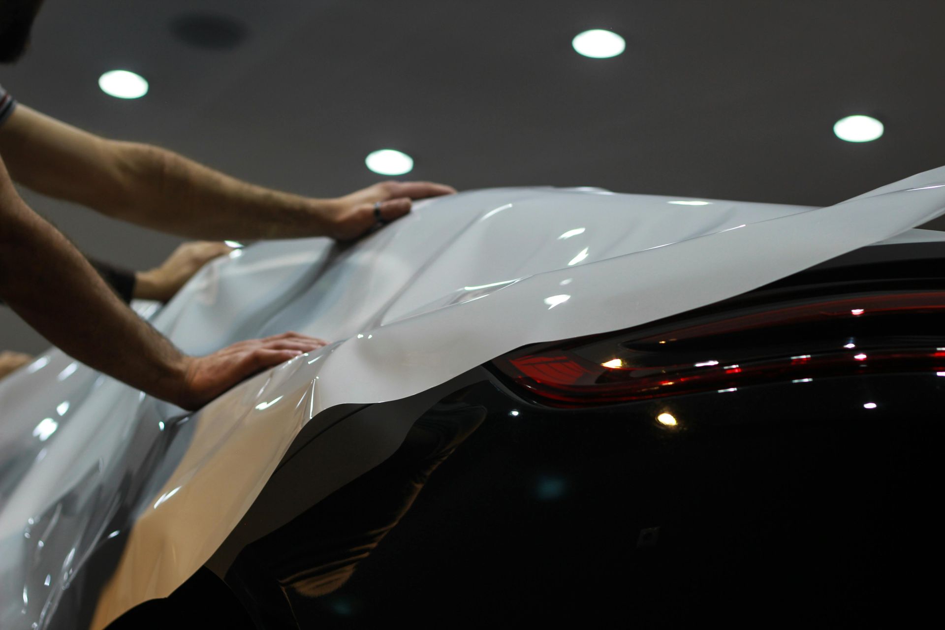Hands applying white vinyl wrap to a black car under bright indoor lights