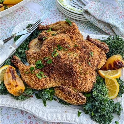 Crispy grilled whole panko chicken on a platter with lemon wedges, herbs, and kale garnish