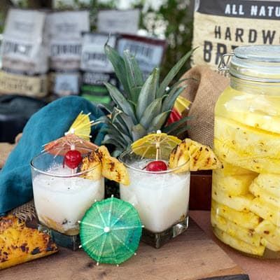 two pina coladas with umbrellas and grilled slices of pineapple garnish, next to a large jar filled with pineapple slices
