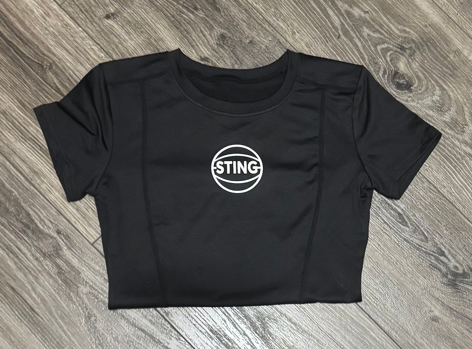 Black Sting T-shirt laid flat on a wood floor, with a white circular logo on the chest