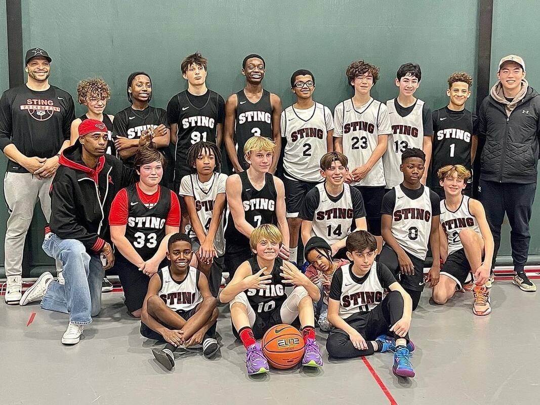 Basketball team posing in a gym, with players in black and white uniforms and a trophy in front