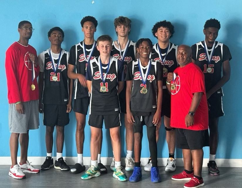 Basketball team posing indoors in black uniforms with medals, standing in front of a blue wall.