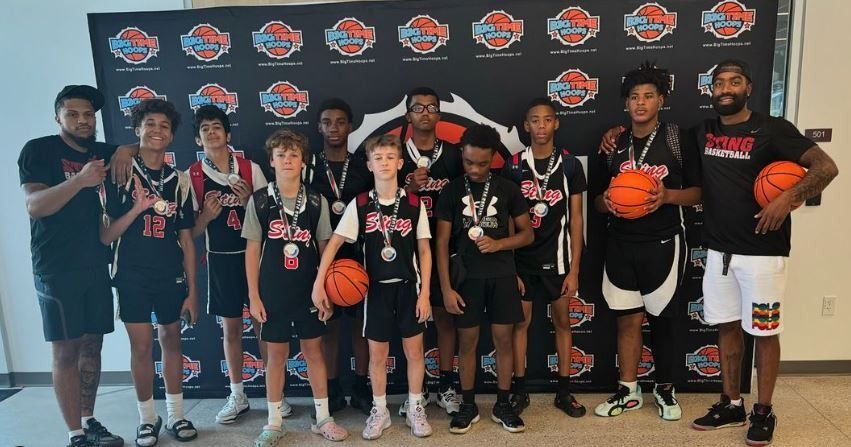 Youth basketball team posing indoors in front of a branded backdrop, holding trophies and basketballs.