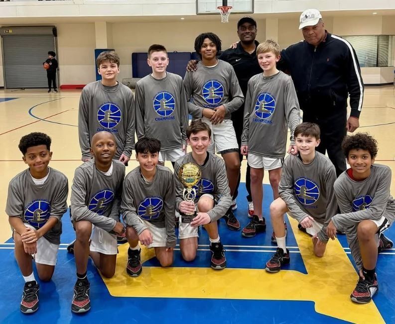 Youth basketball team posing in a gym with trophies and two coaches behind them.