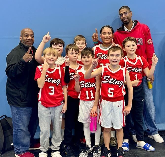 Youth basketball team posing with coaches in red and white uniforms against a blue wall.