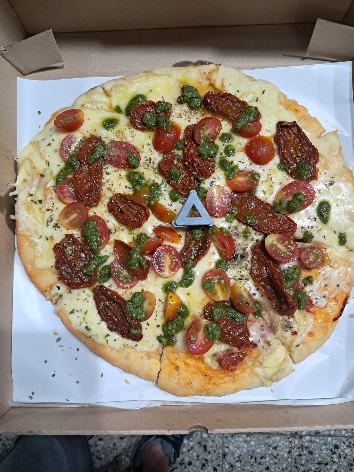 Pizza abierta en caja, cubierta con tomates cherry, hierbas y queso fundido.