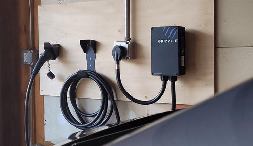 Wall-mounted EV charger with coiled black cable and plug beside a power box on a beige wall