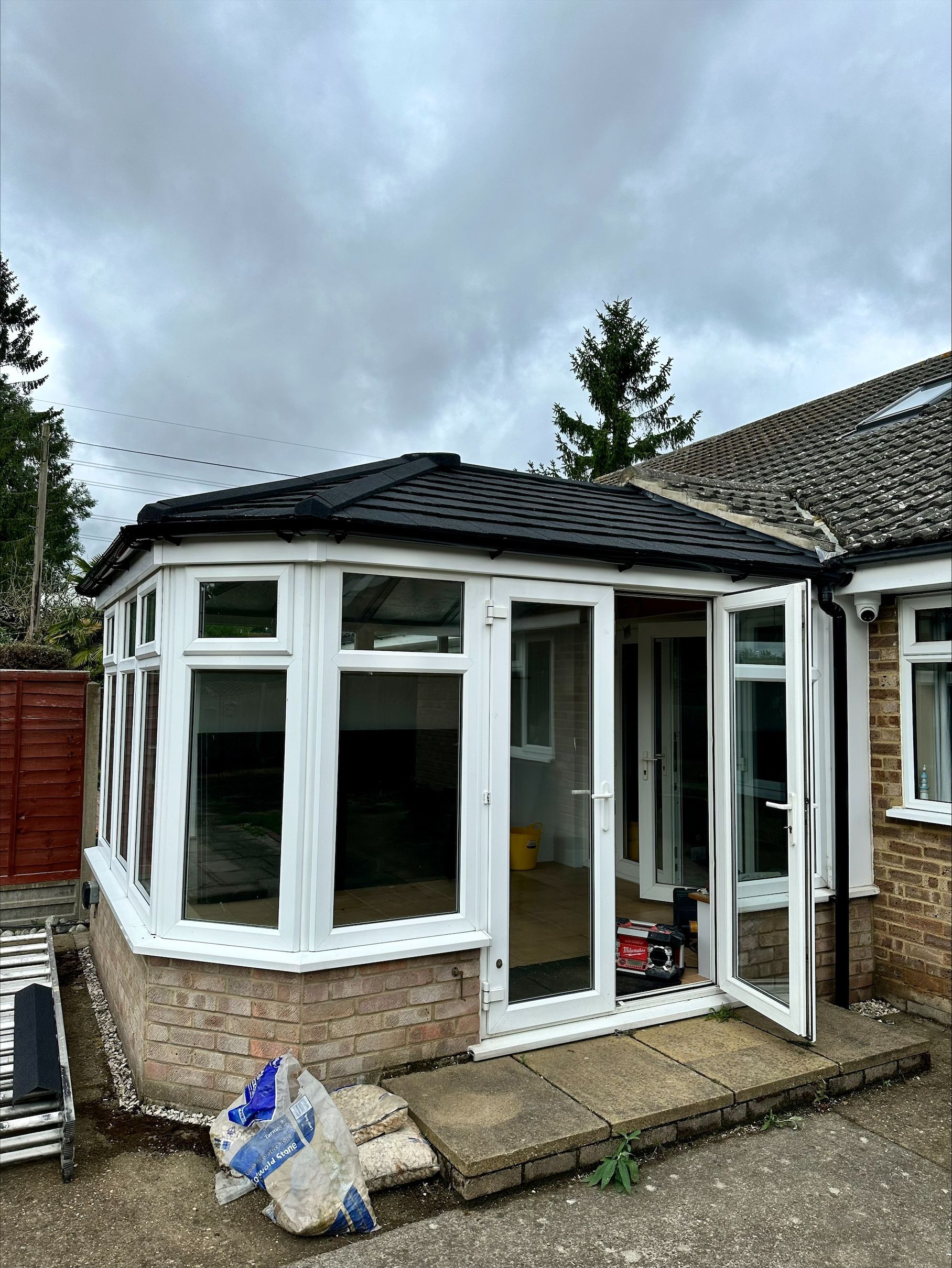 Sunroom addition with white frames, brick base, and dark tiled roof against a cloudy sky.