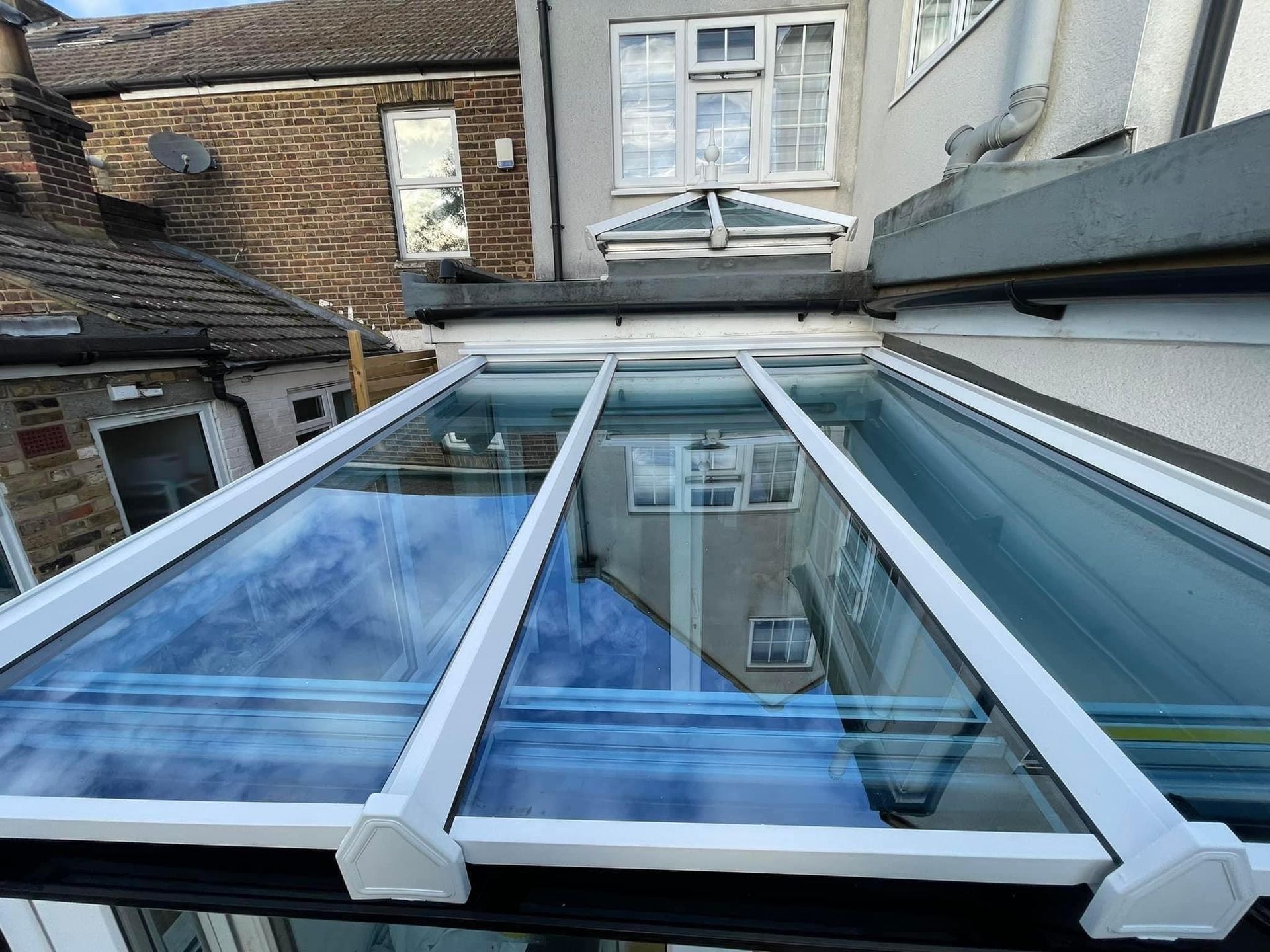 Glass roof of a conservatory with white frames, reflecting the sky and surrounding buildings.