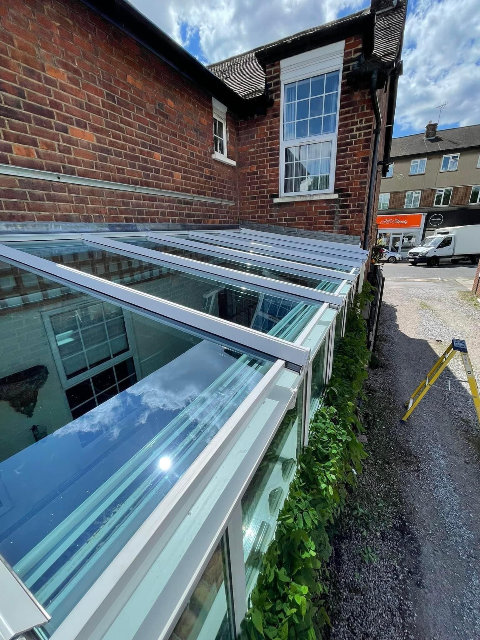 Glass-roofed structure attached to a brick building. Bright sunlight reflects off the glass. Green foliage along the side.