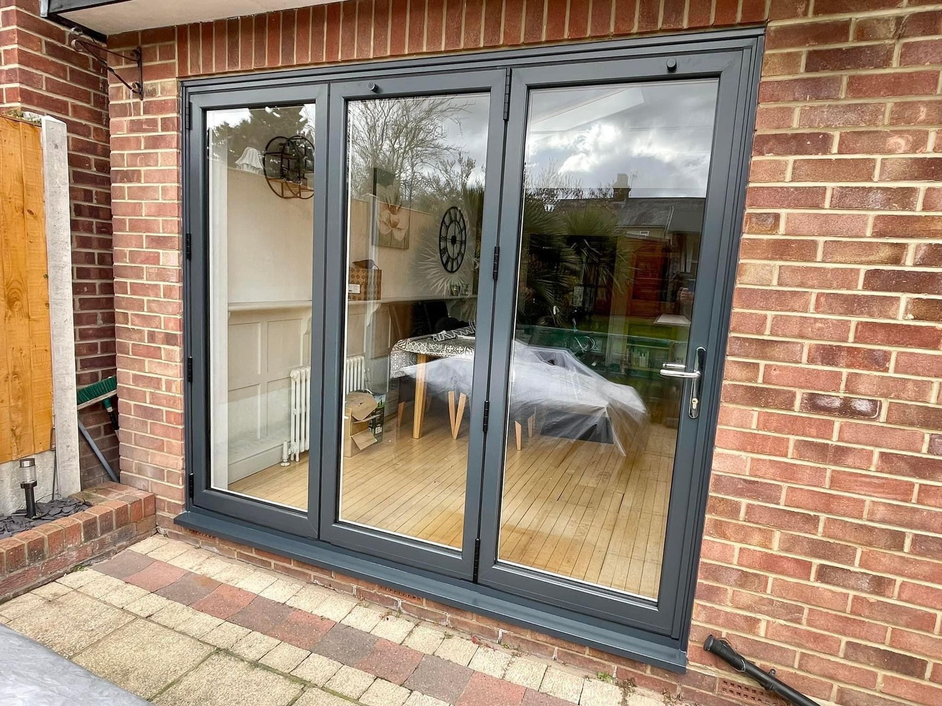 Three-panel gray folding doors on a brick house, reflecting an interior dining area with a table.