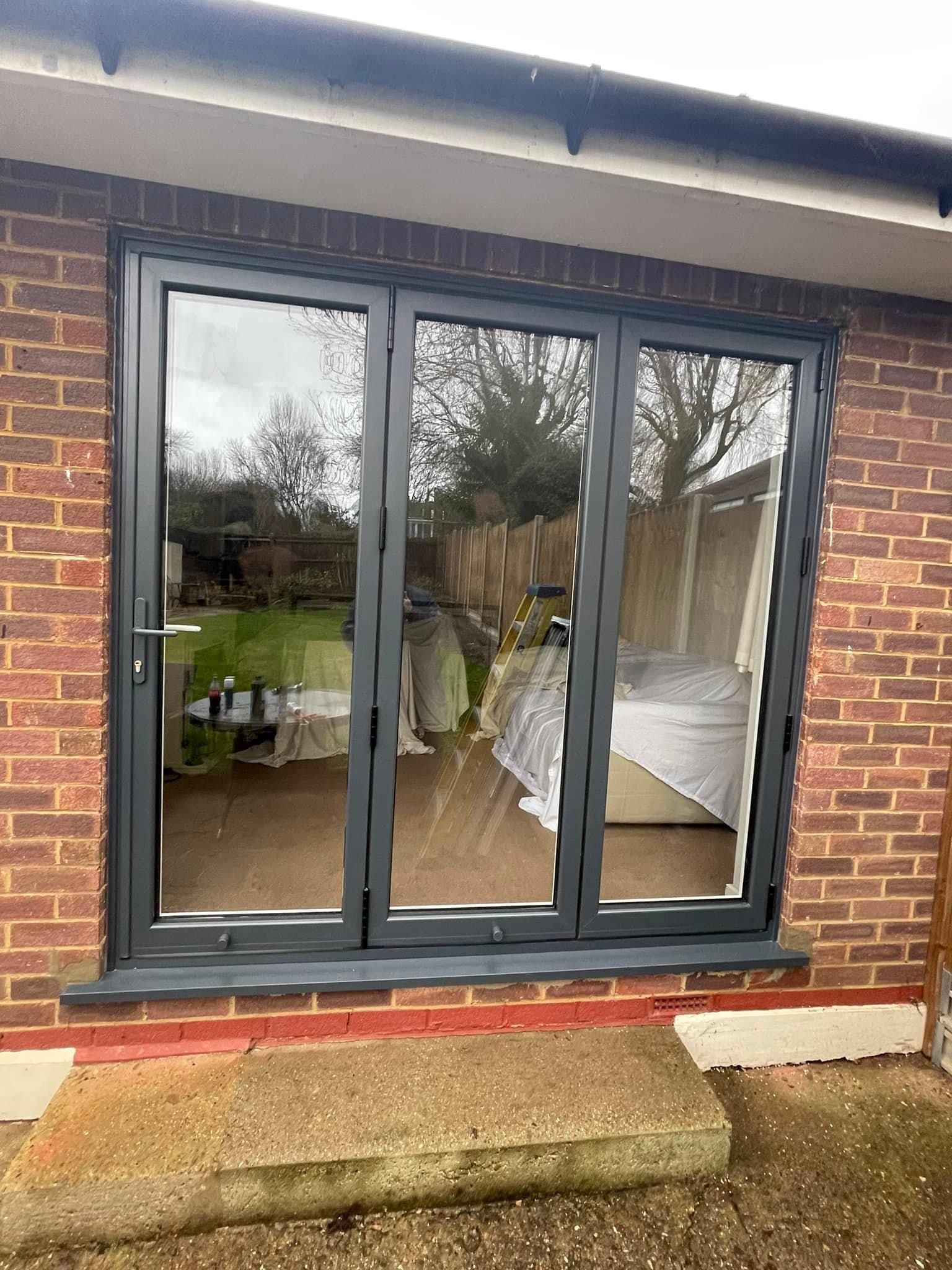 Gray folding doors in a brick wall, reflecting a bedroom interior and outdoor foliage.