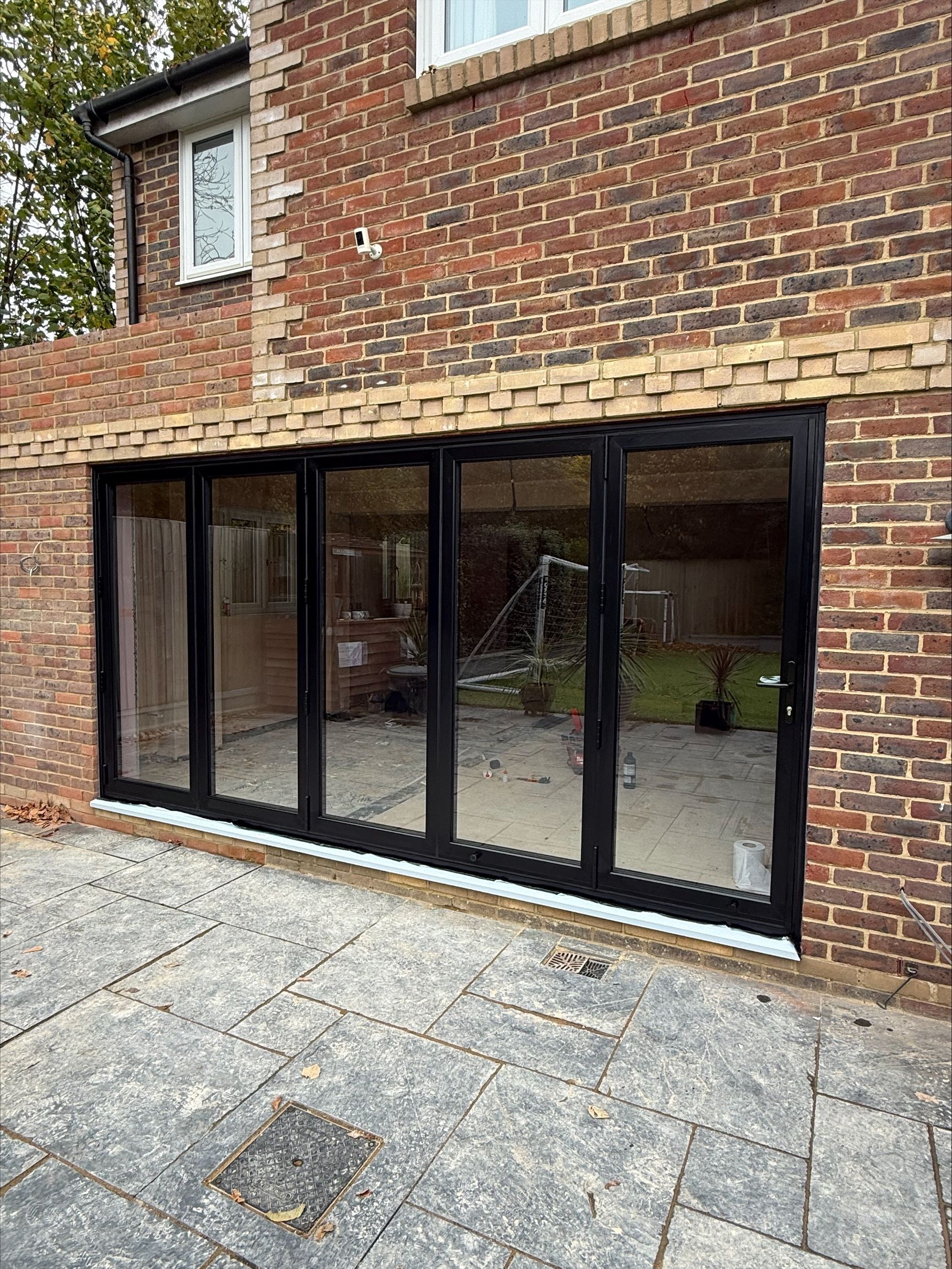 Black framed folding doors in brick building. Paved patio in foreground.