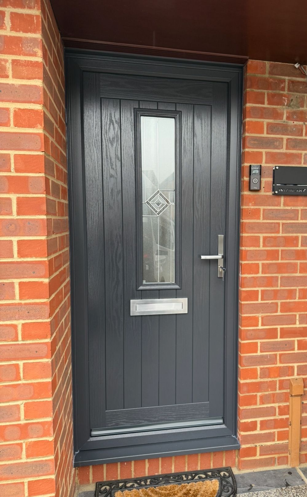 Dark gray front door with vertical grooves, glass panel, and silver hardware set in a red brick wall.