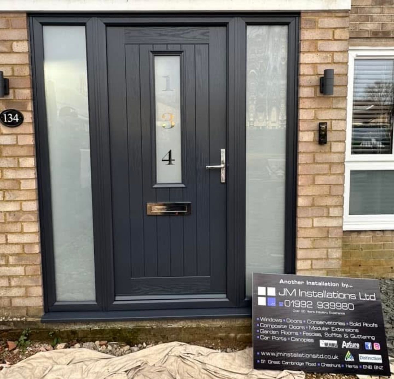 Dark gray front door with sidelights, address 134, sign for JM Installations.