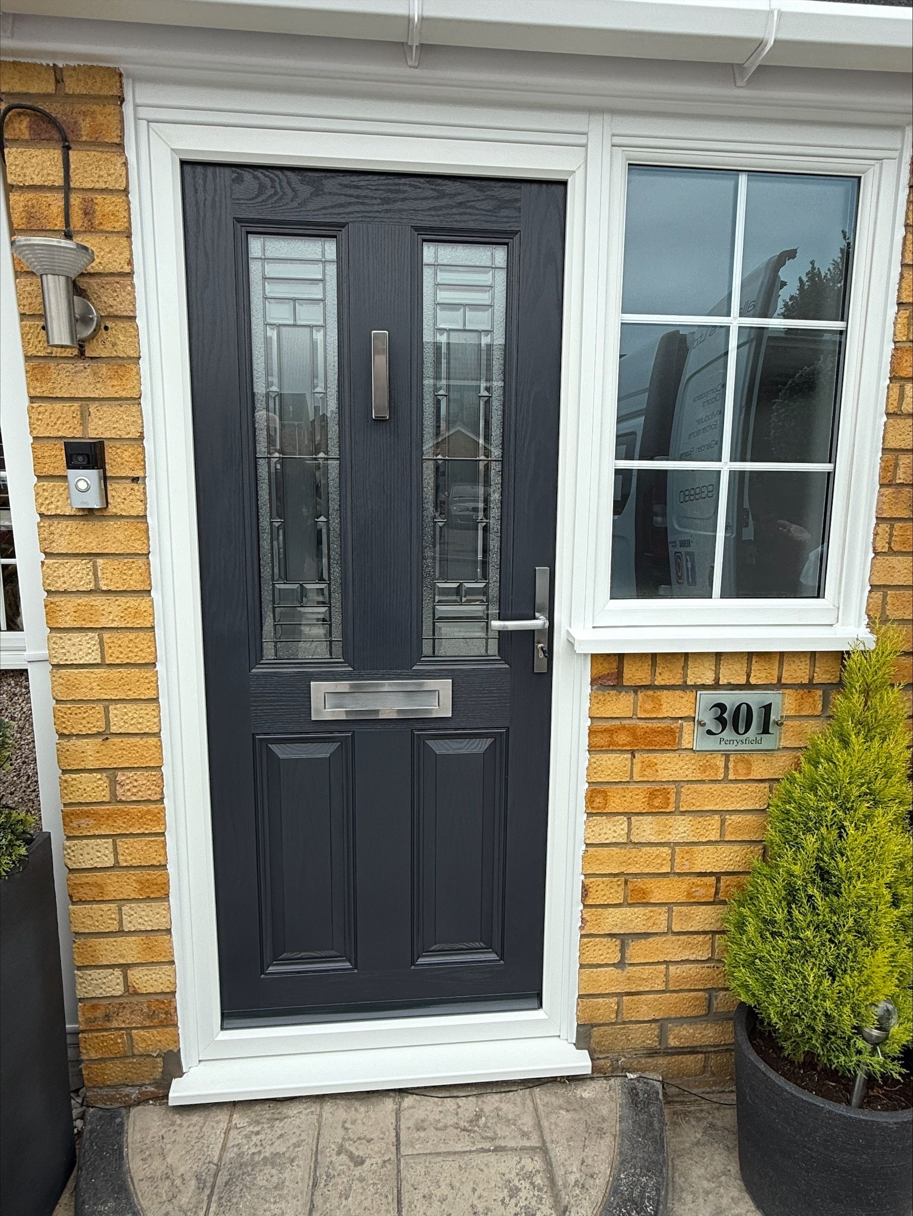 Dark gray front door with glass panels and sidelight, set in a brick facade, white trim, and a small potted plant.