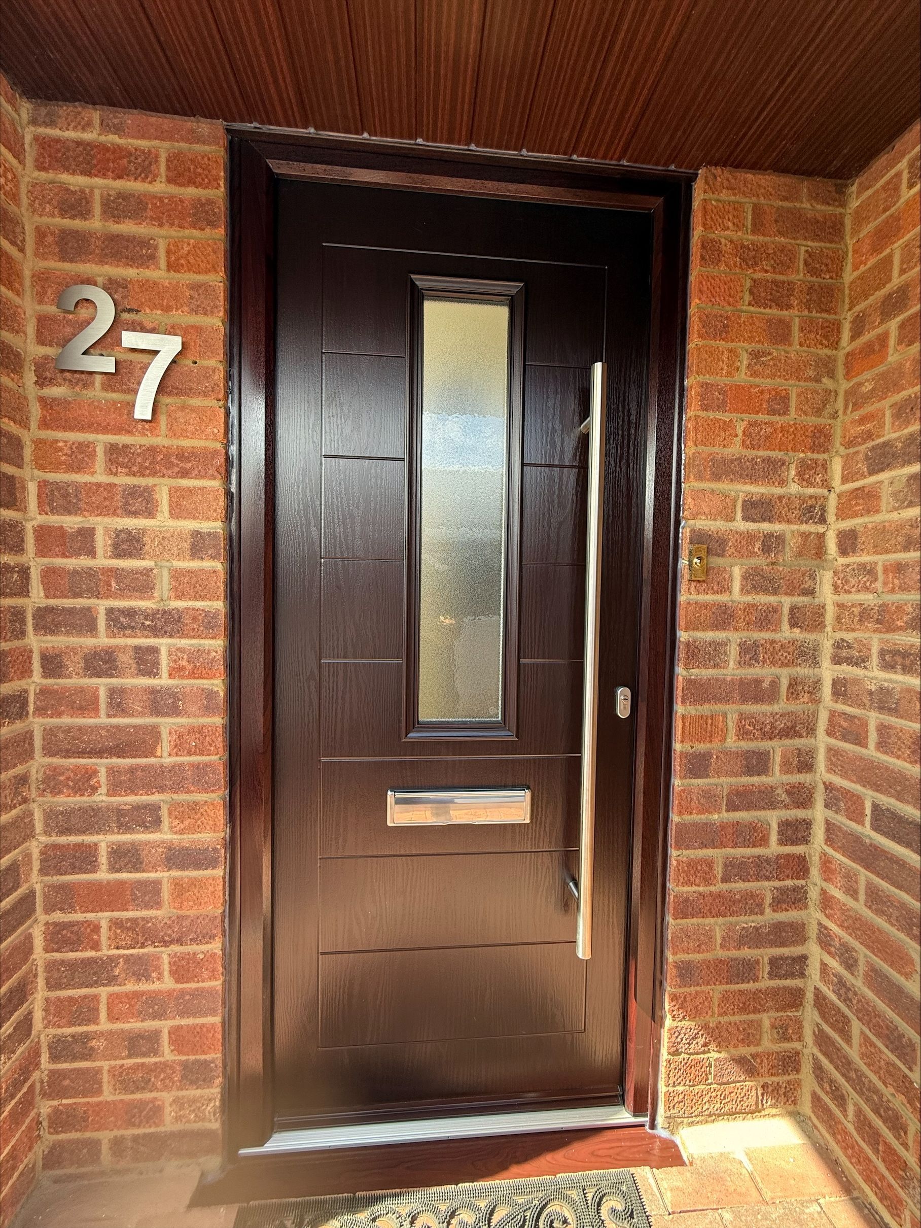 Brown front door with vertical glass panel, brick facade, and silver address number 27.