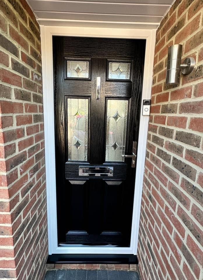 Black front door with decorative glass panels set in brickwork, flanked by a white frame and silver light fixture.