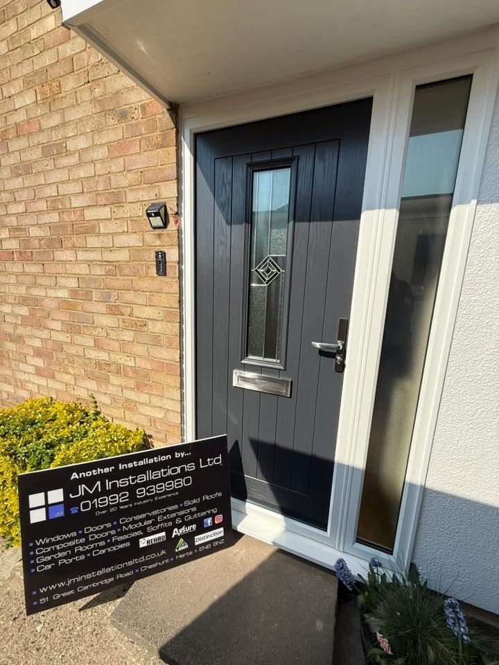 A gray front door with glass panel and white frame, beside a frosted glass window. JVM Installations sign on the ground.