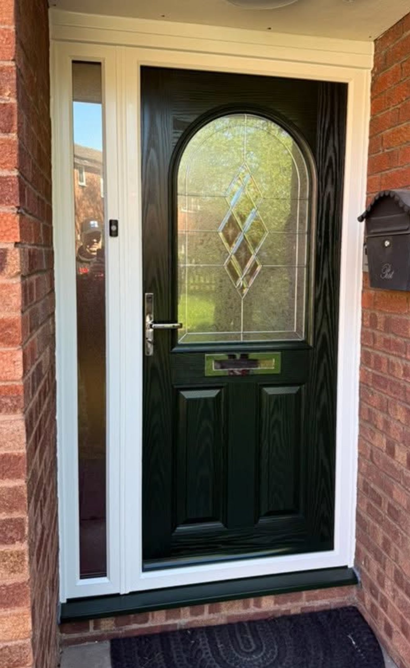 Green front door with arched glass panel and side window, white frame, brick exterior.