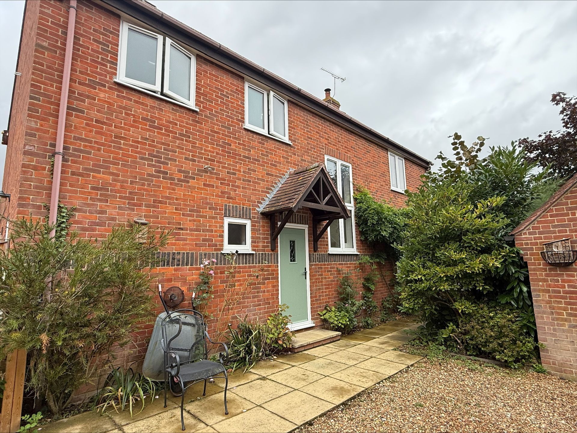 Brick house with green door, small porch, and overgrown front garden. Cloudy sky overhead.
