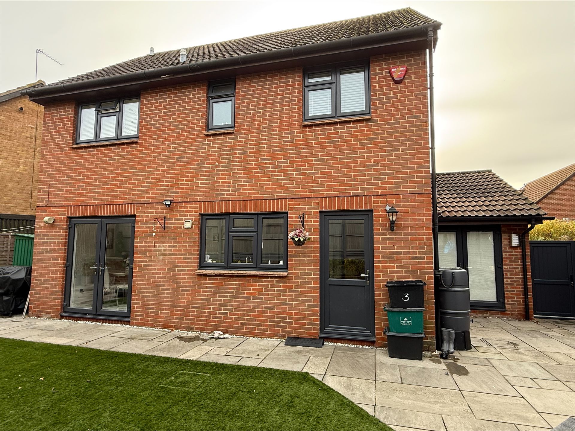 Red brick two-story house with dark gray windows and doors. The yard has paving and a patch of artificial grass.