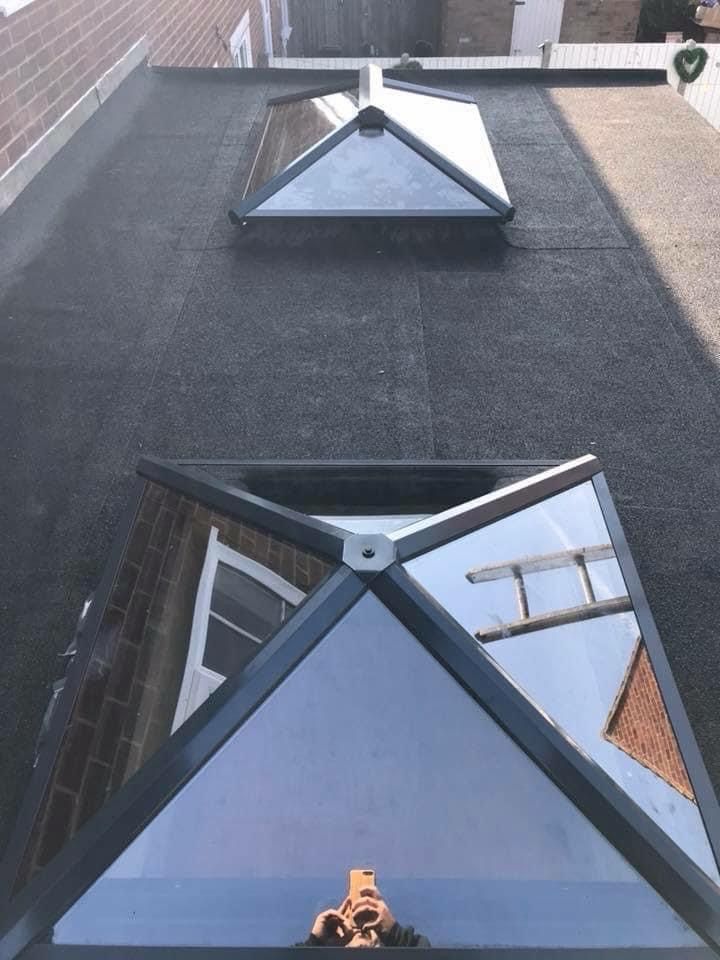 Two triangular skylights on a flat, black roof. Reflection shows a window and ladder.