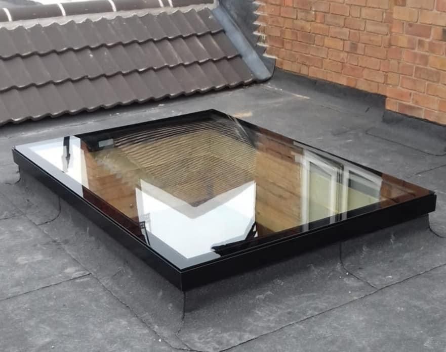 Rectangular skylight on a flat roof with dark shingles and brick wall in the background.