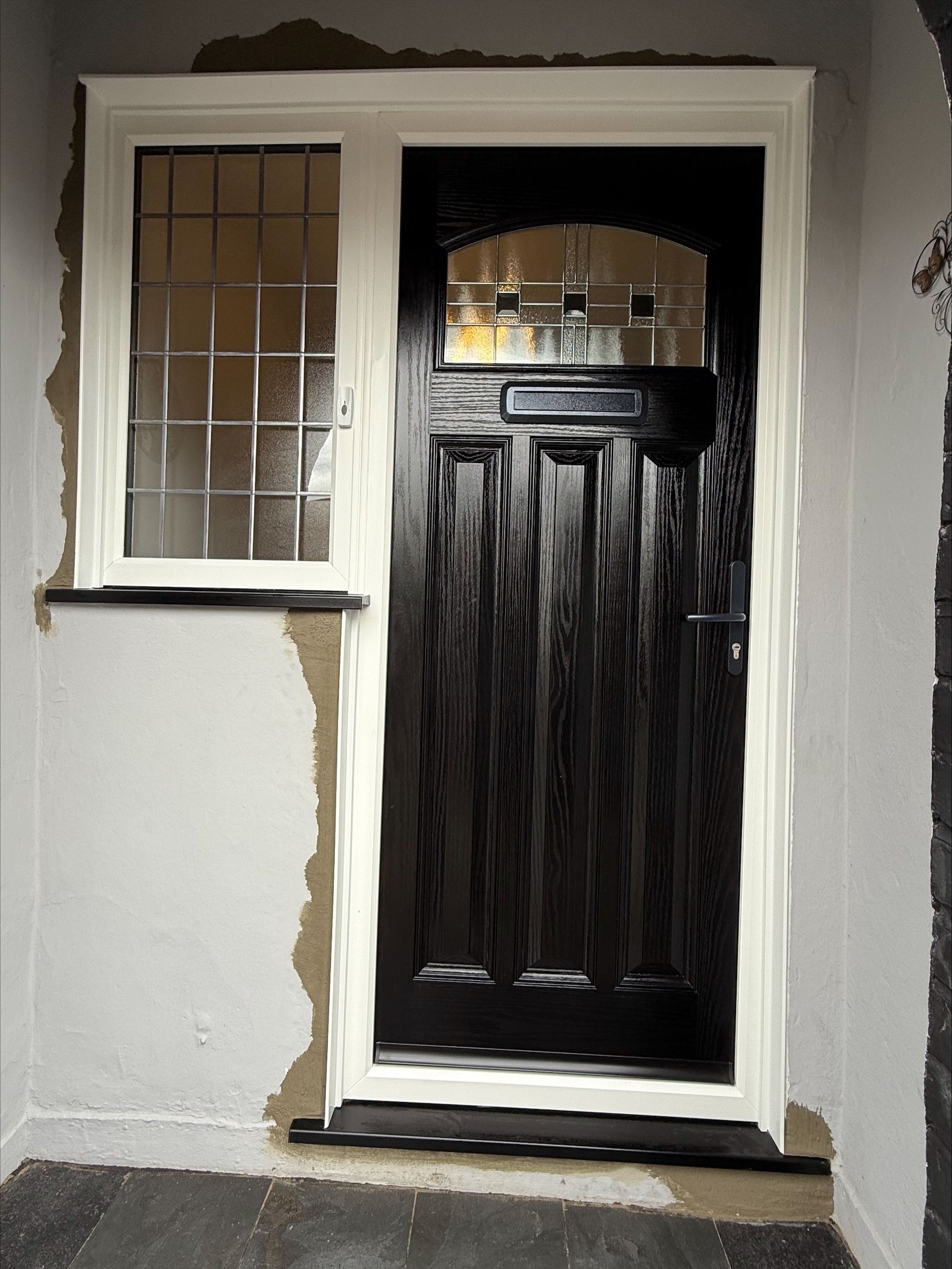 Black front door with sidelight window, framed in white trim, set in a gray exterior wall.