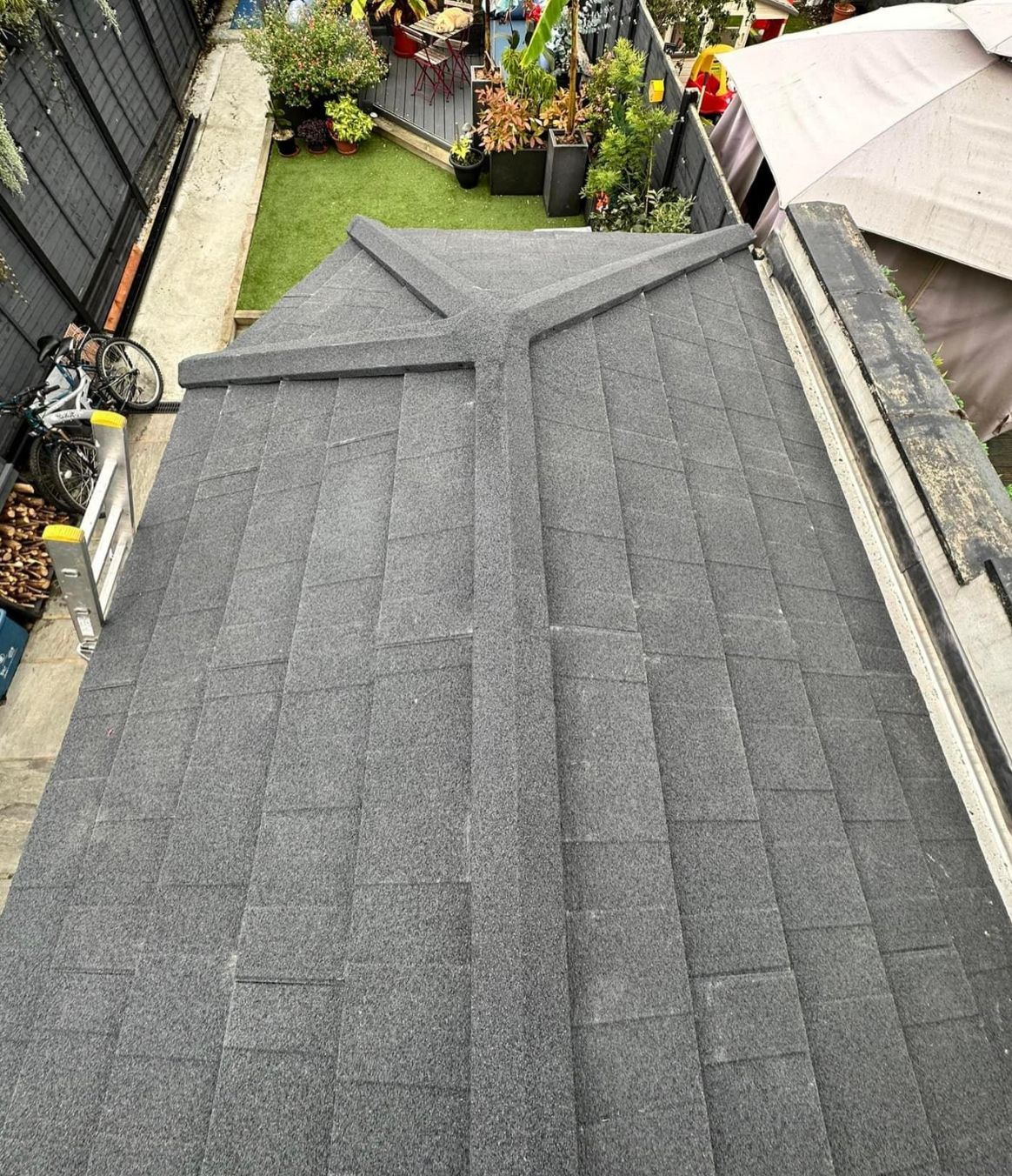 Overhead view of a dark gray shingled roof with a cross-shaped ridge against a garden backdrop.