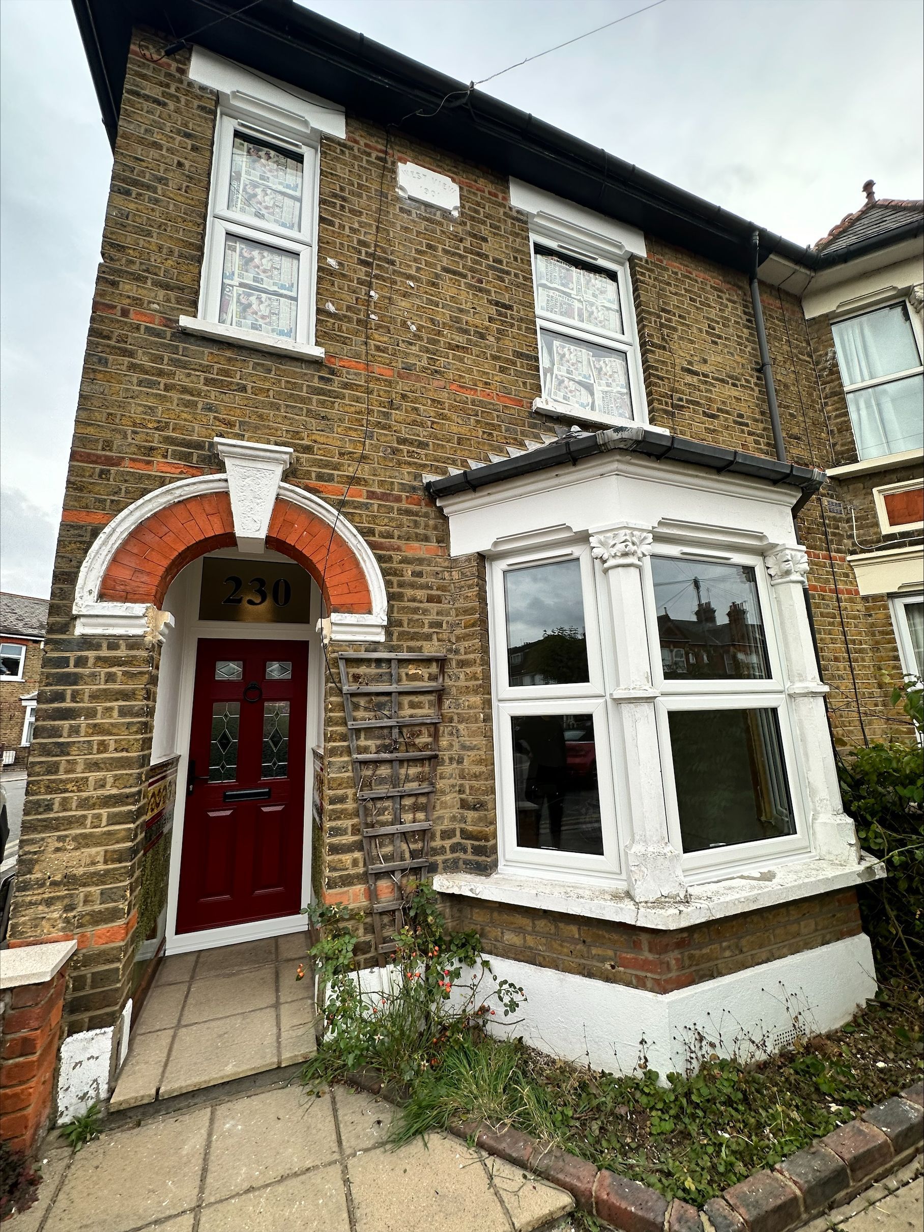 Brick house with red door and white-framed windows, overgrown plants in front.