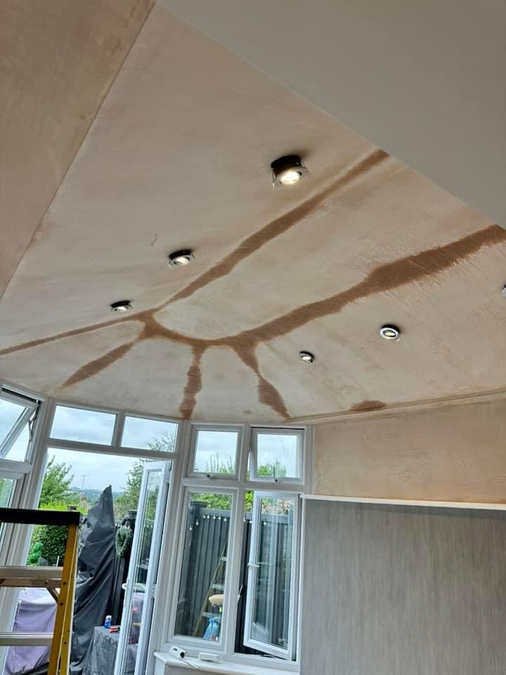 Ceiling of a room with recessed lights and unfinished plasterwork, next to large windows.