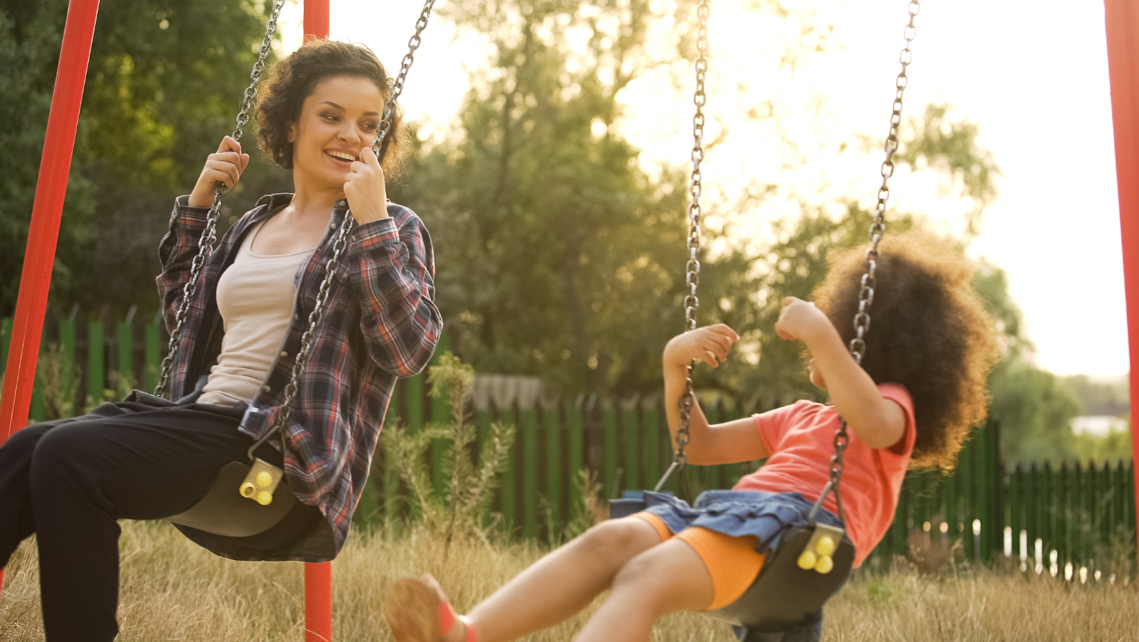 Two children on swings, smiling in a sunny park at golden hour.