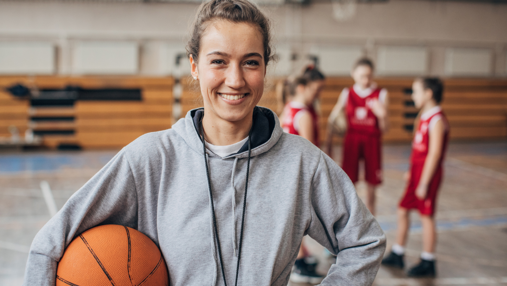 Smiling basketball player in gray hoodie holding a ball on an indoor court, with teammates in the background