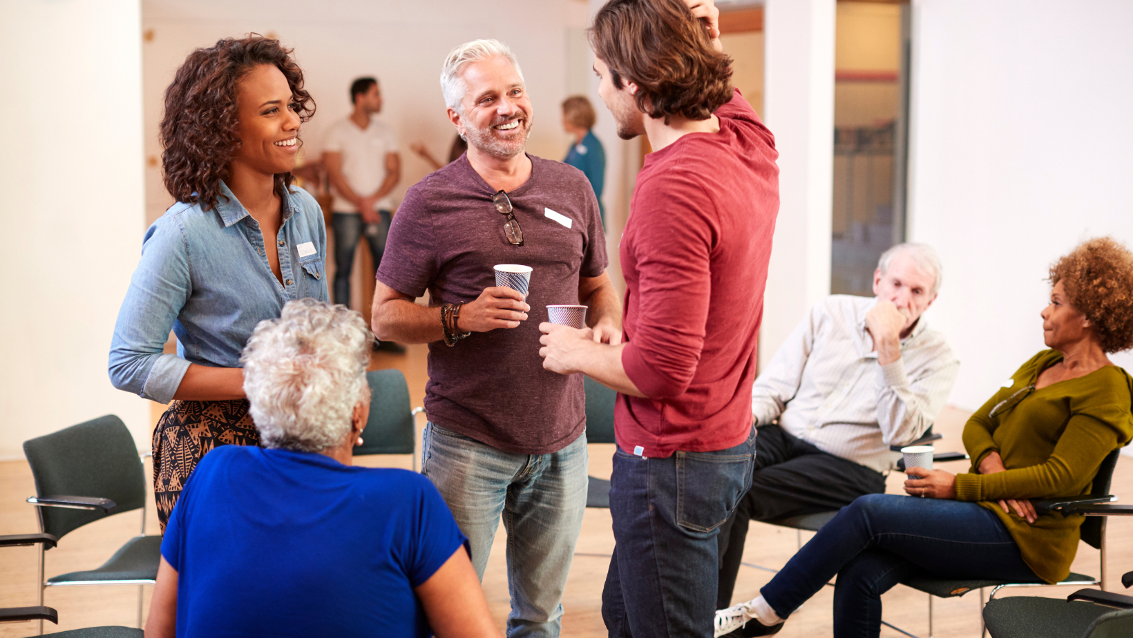 People chatting and laughing in a casual group meeting room, holding drinks and sitting in chairs.