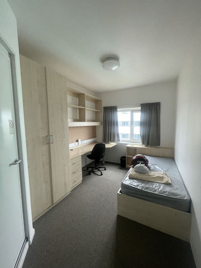 Small student bedroom with bed, desk, chair, shelves, and a window with curtains.
