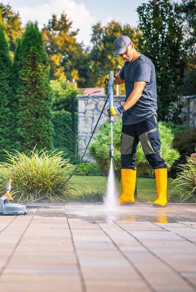 Person pressure-washing a patio in yellow boots and black clothes in a garden.