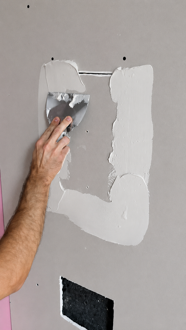 Hand spreading gray plaster over a patch of damaged wall with a putty knife.