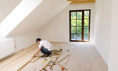 Person installing wooden flooring in a bright empty attic room with a window and tools on the floor