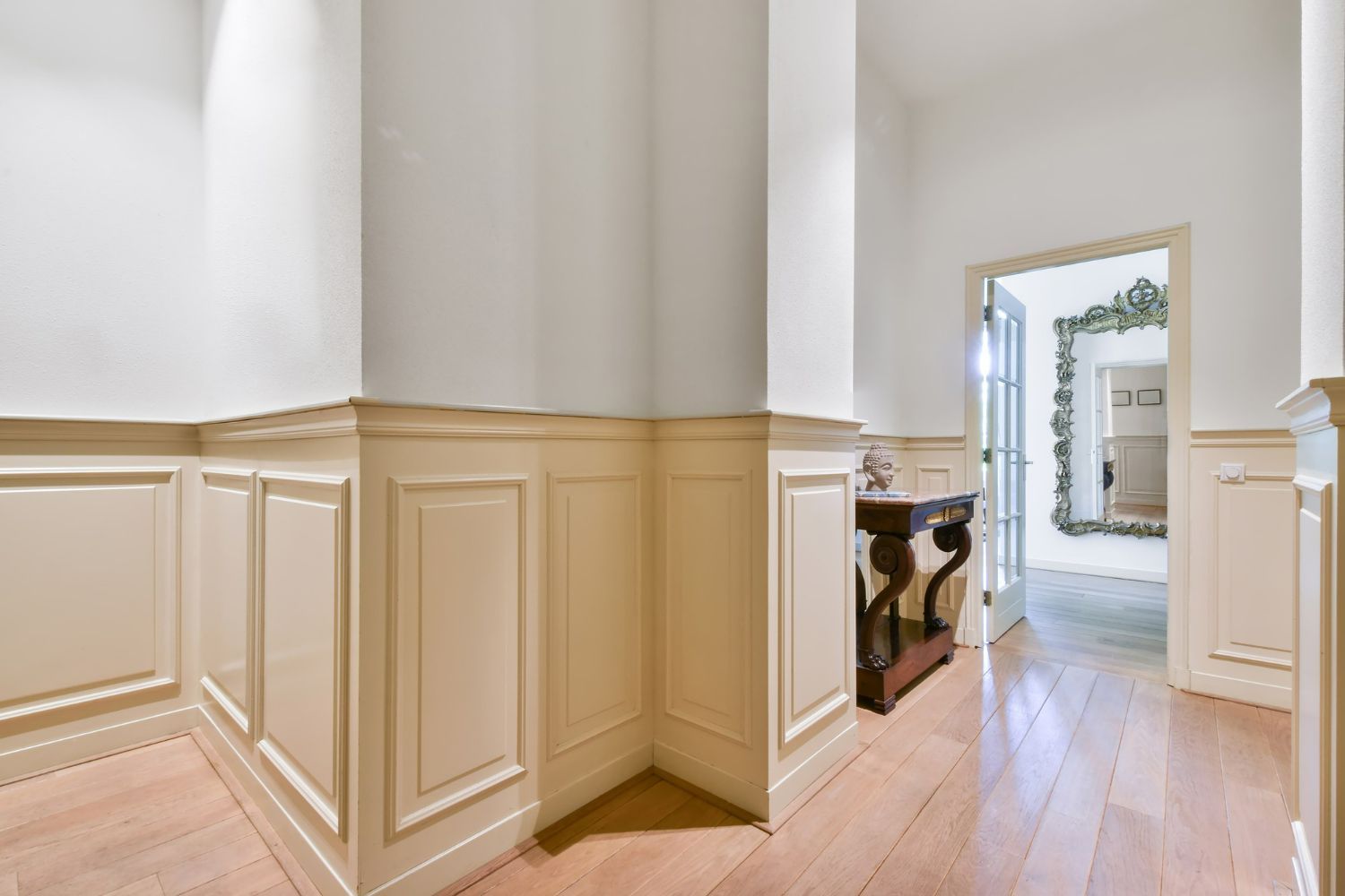 An interior view of a hallway featuring cream-colored wainscoting, white walls, light wood floors, and a doorway beyond.