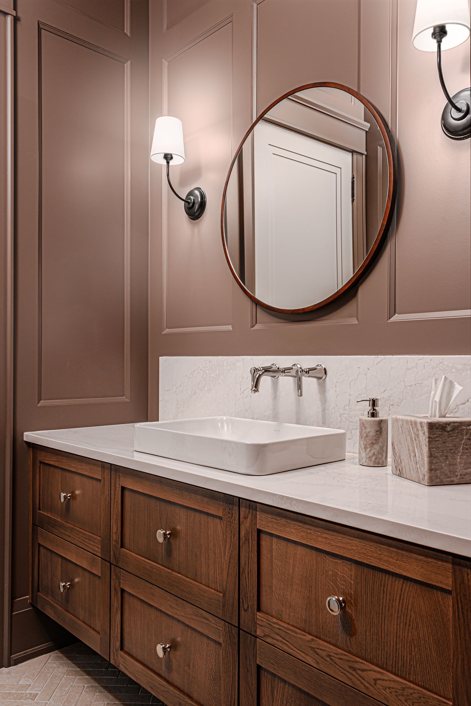 Bathroom vanity with wood cabinets, white sink, round mirror, and wall sconces on a beige wall