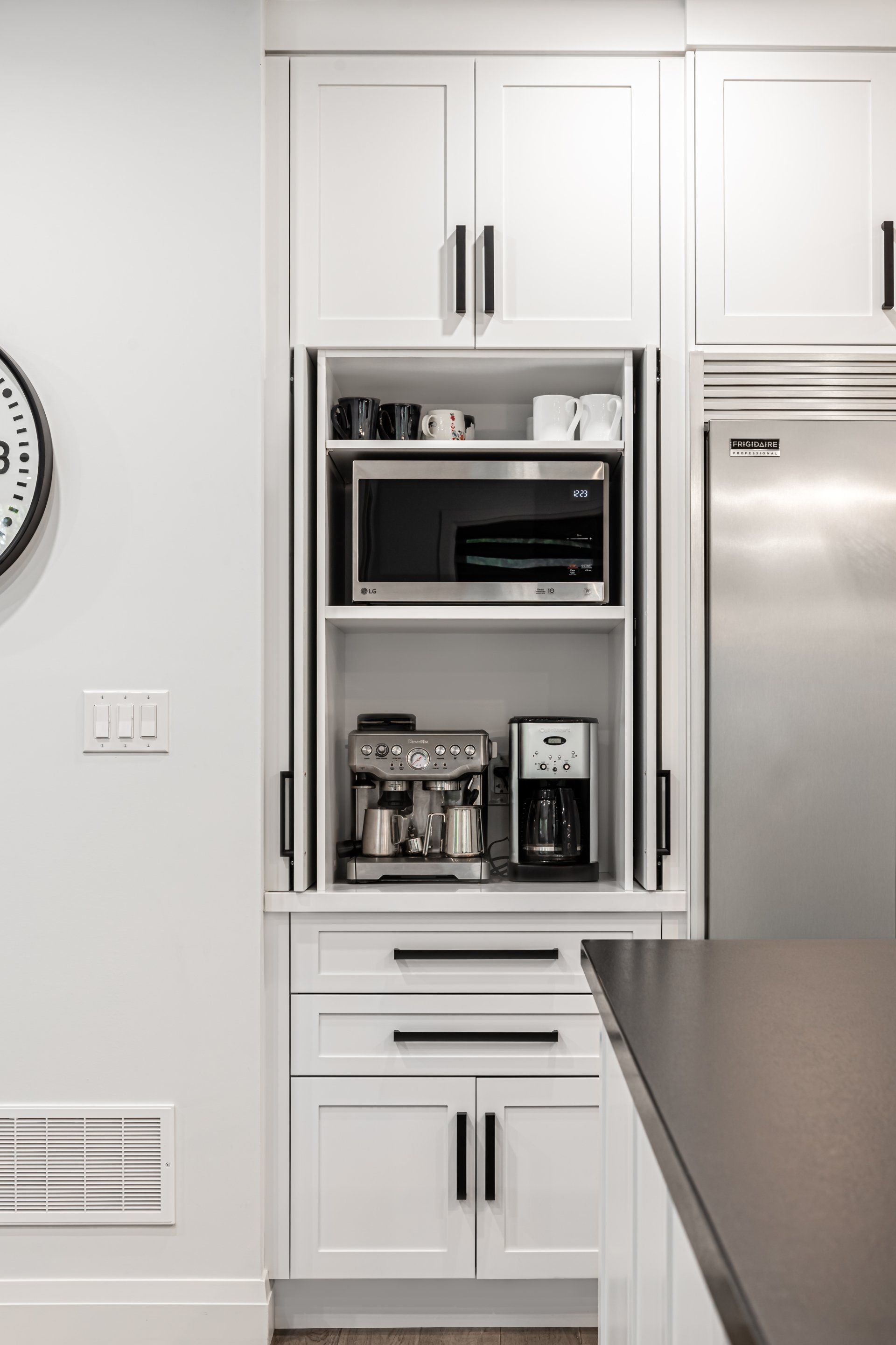 A kitchen with white cabinets , a microwave , a coffee maker , and a clock on the wall.