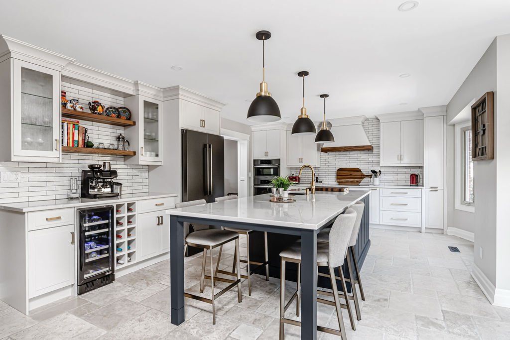 A kitchen with white cabinets , a large island , stools , a refrigerator and a wine cooler.