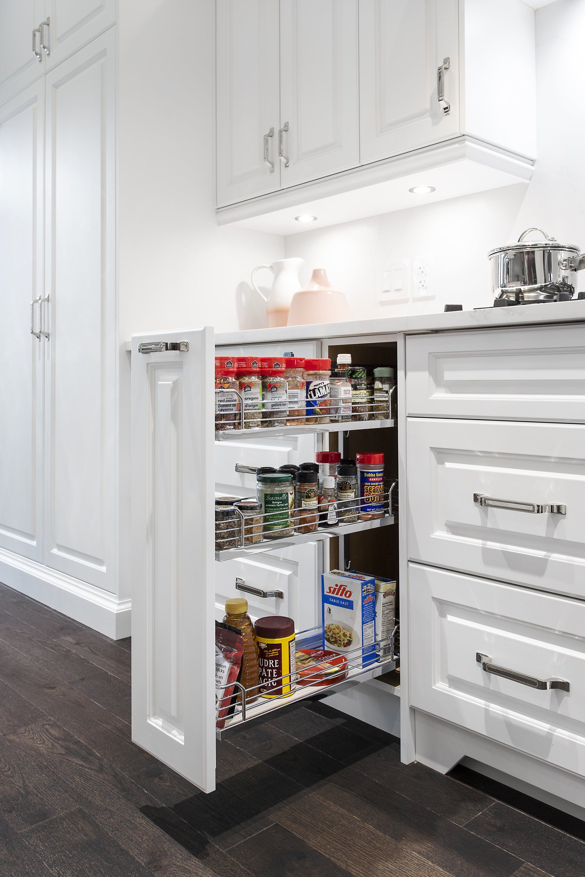 A kitchen with white cabinets and drawers and a pull out pantry filled with food.