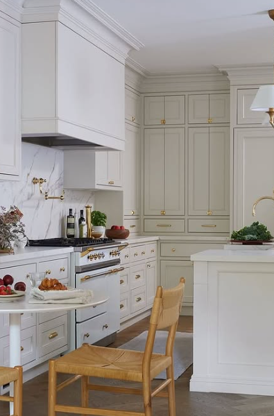 Bright white kitchen with built-in cabinets, wooden chair, stove, and a small island with fruit and greens