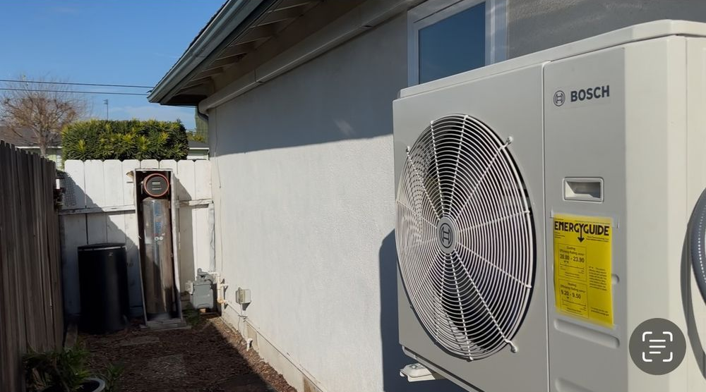 Outdoor HVAC unit beside a house wall, with a narrow side yard and fence in the background.