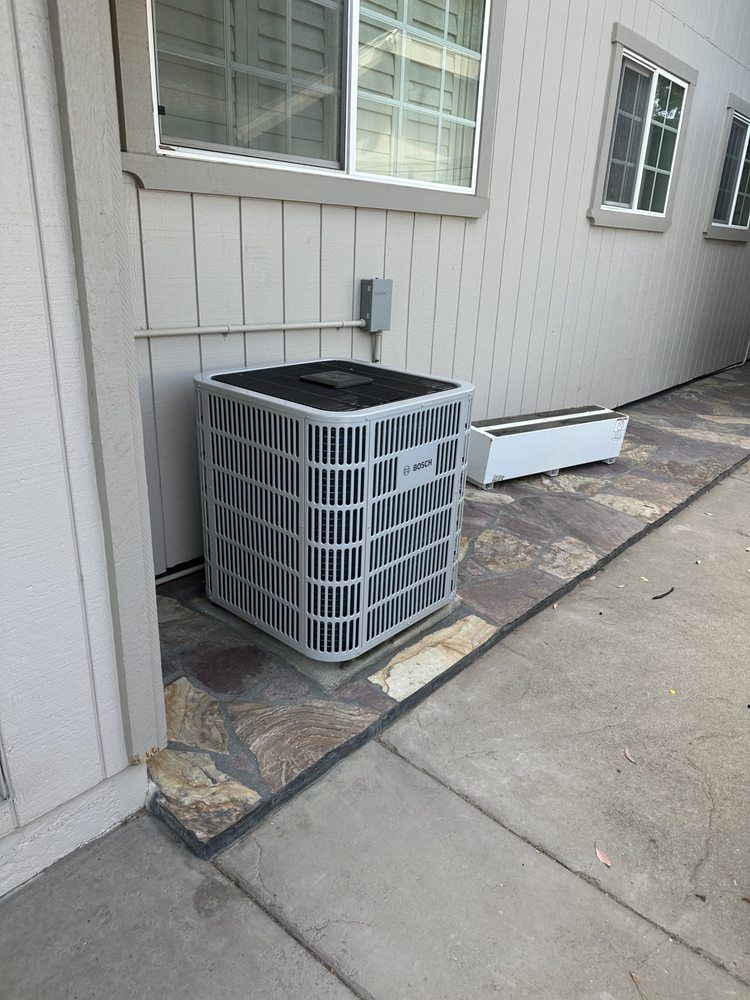 Outdoor HVAC unit beside a house wall, with a window and concrete walkway nearby